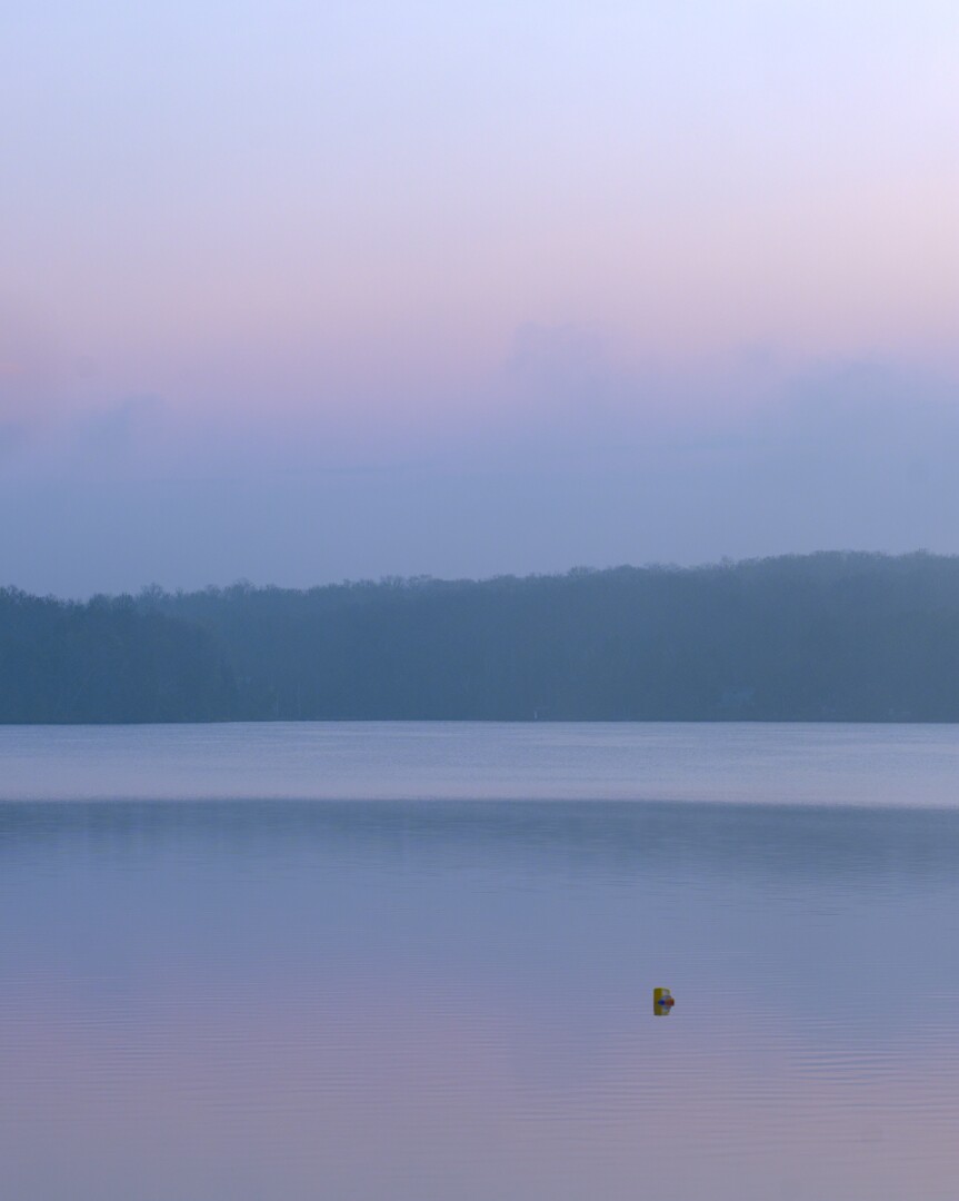 A buoy floating in a calm lake suffused in the pastel light of sunrise