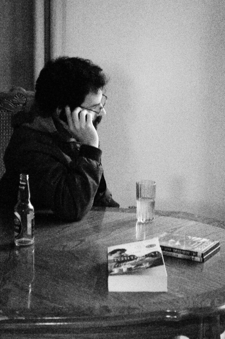 A man on his cellphone at a table with a book and glasses on it. Black and white.